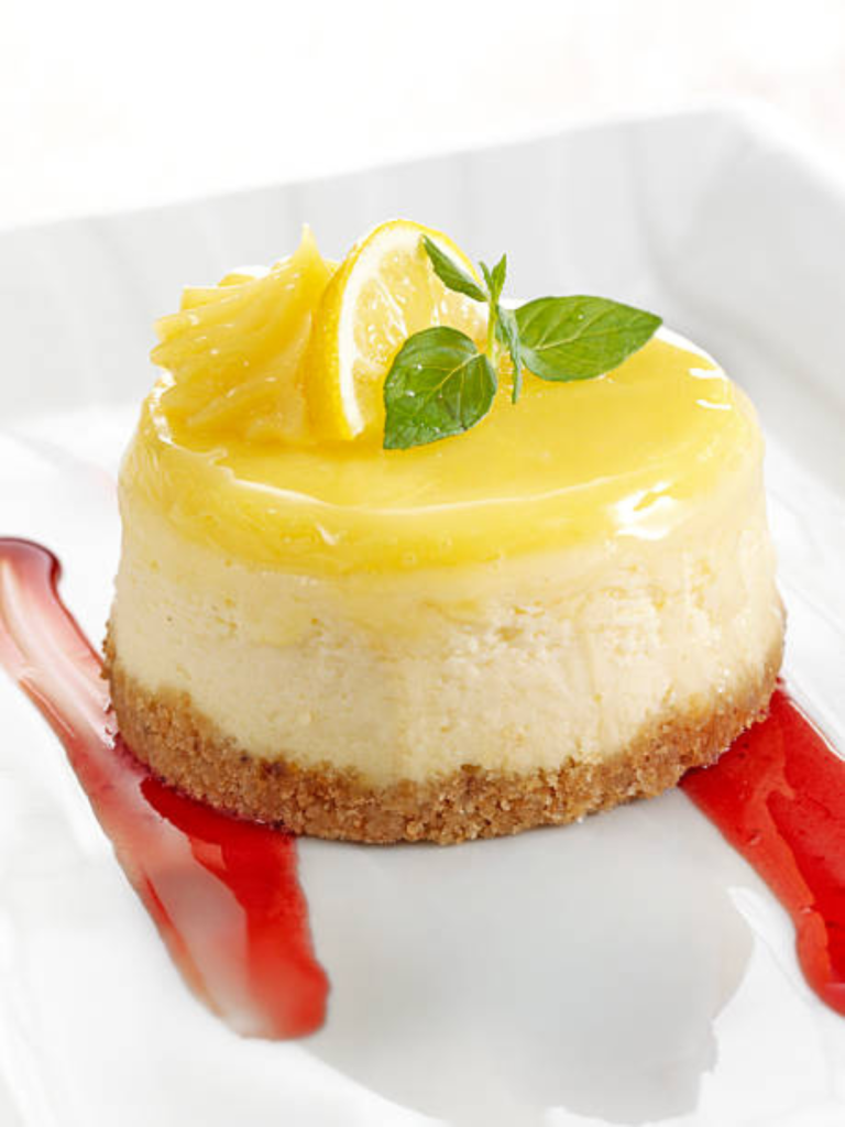 Mini sugar free baked lemon cheesecake topped with lemon curd, a fresh lemon slice, mint leaves, and berry sauce