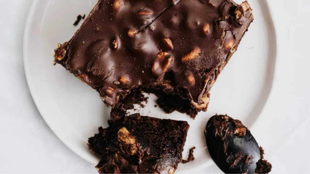 ​A slice of rich sugar-free avocado chocolate brownie topped with shiny chocolate glaze and nuts on a white plate with a black spoon.