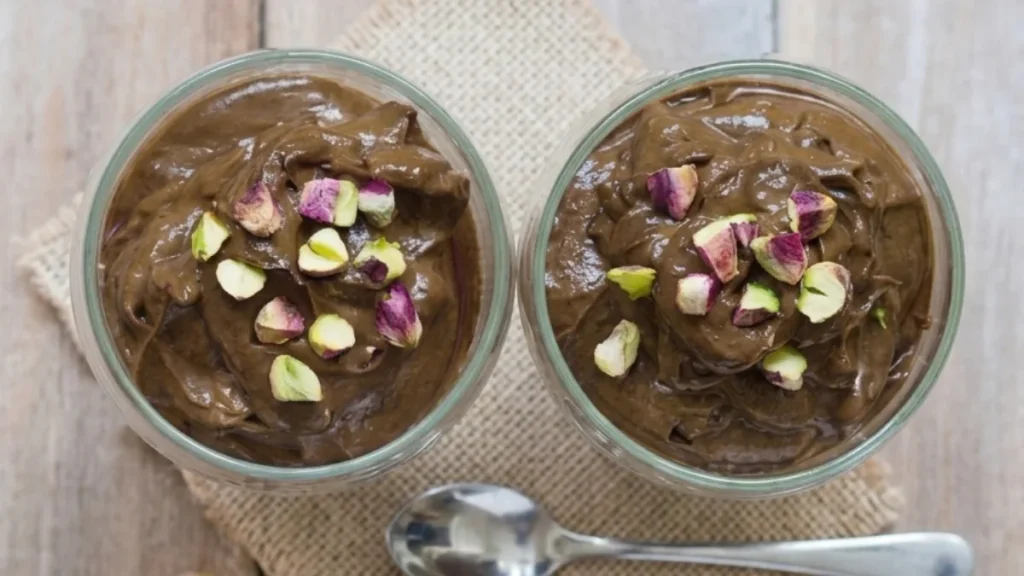 Two glass cups filled with creamy date-sweetened chocolate avocado mousse topped with chopped pistachios on a wooden table with spoons.