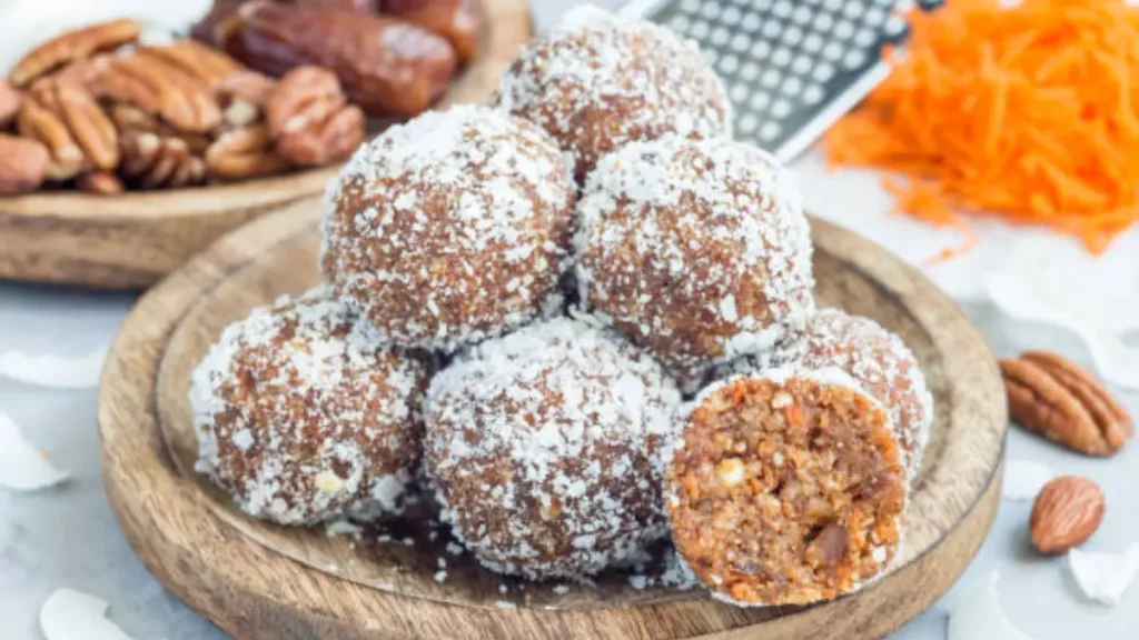 A wooden plate stacked with raw, date-sweetened carrot cake bites rolled in finely shredded unsweetened coconut, with one bite sliced open to reveal the soft, textured carrot and walnut interior.